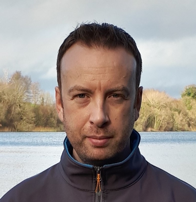 Stephen stands in front of a serene lake, surrounded by trees and a cloudy sky.